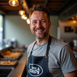 Liam Hudson, fish & chip shop expert, smiling inside a fryer kitchen. Dressed in a branded apron and grey tee, with fryers behind him, ready to serve up crispy goodness.